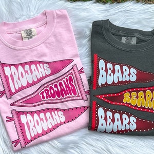 May include: Two t-shirts with pennant designs. The pink shirt has three pennants that say "Trojans" in white. The grey shirt has three pennants that say "Bears" in white.
