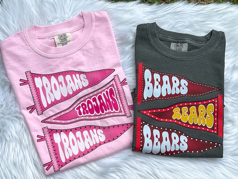 May include: Two t-shirts with pennant designs. The pink shirt has three pennants that say "Trojans" in white. The grey shirt has three pennants that say "Bears" in white.