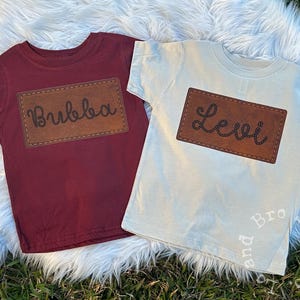 Könnte beinhalten: Zwei T-Shirts für Kinder mit braunen Leder-Patches. Das weinrote Shirt hat den Namen "Bubba" auf dem Patch. Das beige Shirt hat den Namen "Levi" auf dem Patch.