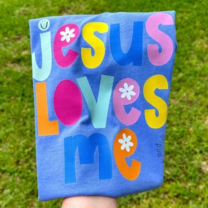 May include: A lilac t-shirt featuring the phrase "JESUS LOVES ME" in vibrant, multi-coloured letters. The design incorporates a smiley face and floral details. The shirt is folded and held against a backdrop of green grass.