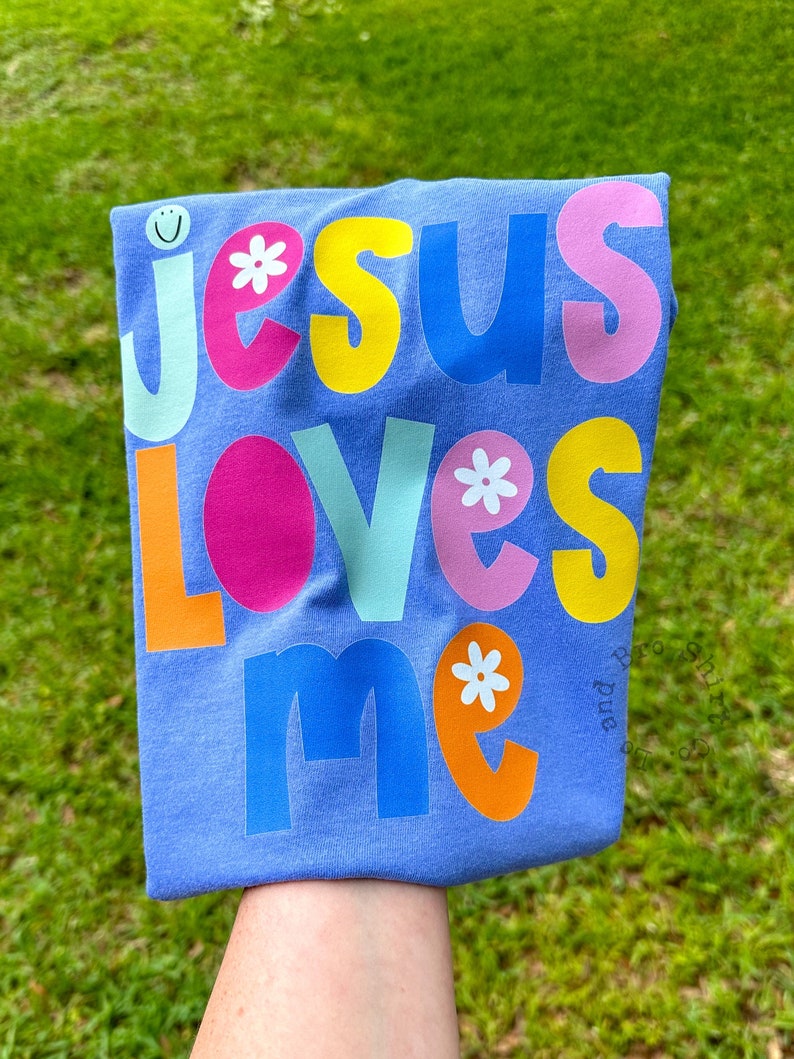May include: A lilac t-shirt featuring the phrase "JESUS LOVES ME" in vibrant, multi-coloured letters. The design incorporates a smiley face and floral details. The shirt is folded and held against a backdrop of green grass.