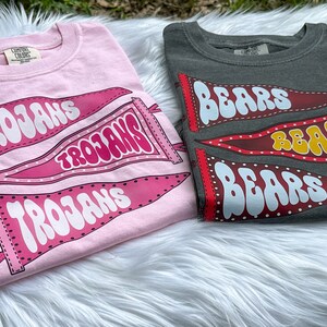 May include: Two t-shirts with different designs. The pink shirt has three pennants with the word "Trojans" on them. The grey shirt has three pennants with the word "Bears" on them.