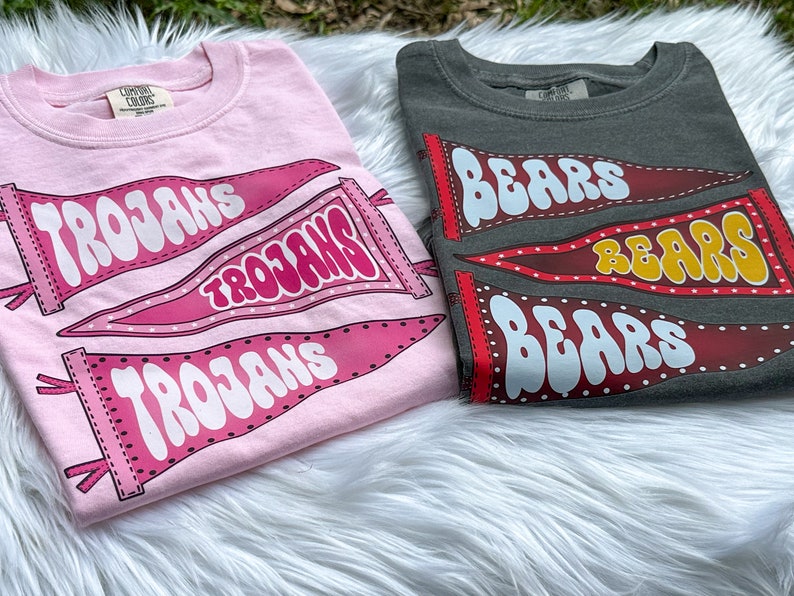 May include: Two t-shirts with different designs. The pink shirt has three pennants with the word "Trojans" on them. The grey shirt has three pennants with the word "Bears" on them.