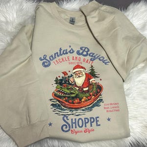 May include: A beige long-sleeve t-shirt with a graphic print of Santa Claus riding a gator with a red boat filled with presents. The text "Santa's Bayou Tackle and Bait Since 1979" is printed above the graphic. The text "SHOPPE Cajun Style" is printed below the graphic.