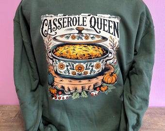 Puede incluir: Sudadera verde con un gráfico de una fuente para hornear y el texto "Casserole Queen". El diseño incluye calabazas y detalles florales. La sudadera se muestra en un maniquí.