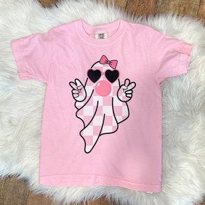 以下が含まれることがあります： ピンクのTシャツに、チェック柄の幽霊がサングラスとリボンをつけています。幽霊はピースサインを2つ上げています。