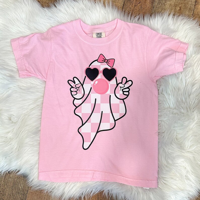 以下が含まれることがあります： ピンクのTシャツに、チェック柄の幽霊がサングラスとリボンをつけています。幽霊はピースサインを2つ上げています。