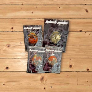 Könnte beinhalten: Vier Black Moon-Emaille-Pins in einzelnen durchsichtigen Plastikverpackungen. Die Pins zeigen verschiedene Designs, darunter einen rot-weißen Roboter, einen gold-weißen Ritter, einen rot-weißen Roboter mit einer Krone und einen rot-weißen Roboter mit einer Krone und einem Schwert.