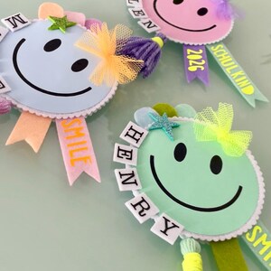 Könnte beinhalten: Drei bunte Smiley-Abzeichen mit Bändern. Die Abzeichen sind hellblau, rosa und grün, jedes mit einem schwarzen Smiley. Die Abzeichen haben dekorative Elemente wie Schleifen, Sterne und Quasten. Ein Band sagt "SMILE", ein anderes "SCHULKIND 2026" und ein weiteres "HENRY".