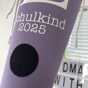 Könnte beinhalten: Ein lilafarbener Turnbeutel mit weißem Text "Schulkind 2025". Der Beutel hat einen schwarzen runden Aufnäher und weiße Kordelzüge. Ein praktischer Artikel für die Schule.