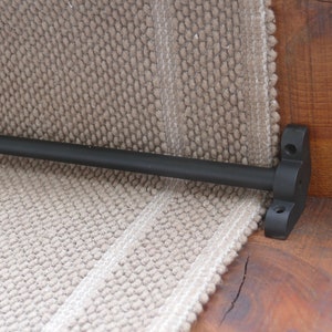 May include: Black metal stair rod with a black metal bracket mounted to a wooden surface. The stair rod is attached to a beige carpet runner.