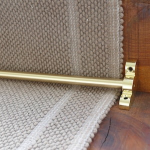 May include: A gold metal rod with a decorative end cap is attached to a beige carpet runner. The rod is used to hold the runner in place on stairs.