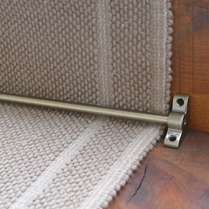 May include: A close-up of a brass stair rod with a textured finish. The rod is attached to a wooden stair tread with a metal bracket.
