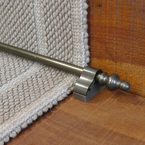 May include: A metal stair rod with a decorative finial in an antique brass finish. The rod is mounted to a wooden stair tread and is holding a beige carpet runner in place.