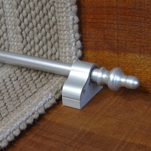 May include: Silver stair rod with a decorative finial. The rod is mounted to a wooden stair tread with a silver bracket.