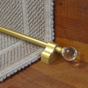 May include: Gold metal stair rod with a crystal finial. The rod is mounted to a wooden surface and a beige carpet is visible in the background.