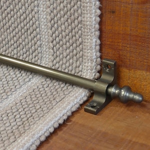 May include: A close-up of a bronze stair rod with a decorative finial. The rod is mounted to a wooden surface and is attached to a beige carpet runner.