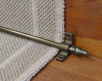 May include: A close-up of a bronze stair rod with a decorative finial. The rod is mounted to a wooden surface and is attached to a beige carpet runner.