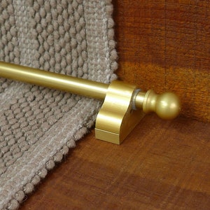 May include: A gold metal stair rod with a round knob on the end. The rod is attached to a wall with a bracket.
