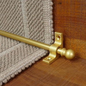 May include: A gold metal stair rod with a round finial. The rod is attached to a wall with a gold metal bracket.