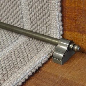 May include: A close-up of a brass stair rod with a decorative end cap. The rod is mounted to a wooden stair tread and is holding a beige carpet runner in place.