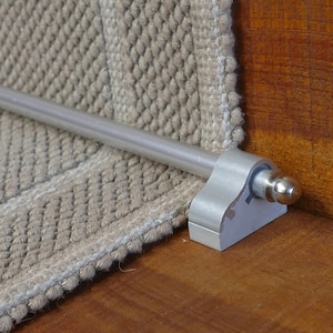 May include: A silver stair rod with a round, silver knob at the end. The rod is attached to a silver bracket that is mounted to a wooden surface. The rod is positioned over a beige carpet.