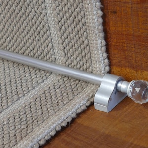 May include: A silver stair rod with a crystal finial. The rod is mounted to a wooden stair tread and is attached to a beige carpet runner.