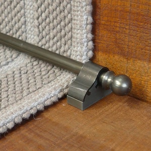 May include: A silver metal stair rod with a ball finial. The rod is mounted to a wooden stair tread.
