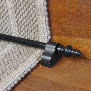 May include: Black metal stair rod with a decorative finial. The rod is mounted to a beige carpet runner.