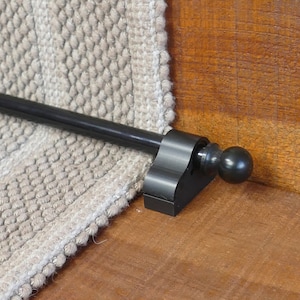 May include: Black metal stair rod with a round finial. The rod is mounted to a wooden stair tread and is holding a beige carpet runner in place.