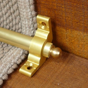 May include: A gold-toned metal stair rod with a ball finial and a decorative bracket. The rod is attached to a beige carpet runner.