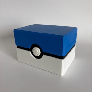 May include: A rectangular storage box designed with a blue top and a white bottom, separated by a black horizontal band. A circular white button is centered on the front, mimicking the appearance of a Great Ball from the Pokemon franchise.
