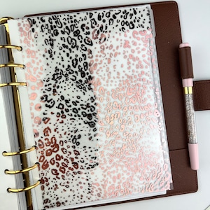 May include: A planner with gold rings, a brown cover, and a clear overlay featuring a leopard print in black and rose gold. A pen with a silver glitter barrel and pink accents rests on the cover.