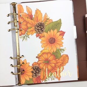 May include: A planner insert featuring a fall-themed design with sunflowers, pumpkins, and leaves in shades of orange, yellow, and green. The name "Heather" is written in gold script. The planner has gold rings and a brown cover.