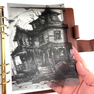 Peut inclure: Illustration en noir et blanc d'une maison hantée avec une pleine lune en arrière-plan. L'image est imprimée sur une feuille transparente, tenue dans un agenda en cuir marron. La maison a un porche et une apparence penchée.
