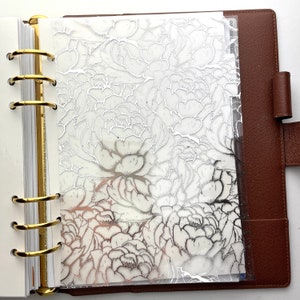 May include: A clear planner divider with a silver floral design. The divider is inserted into a brown leather planner with gold rings. The floral pattern is a detailed outline of peony flowers.