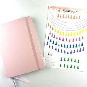 Puede incluir: Un diario rosa claro con cierre elástico y cinta de marcador junto a una hoja de pegatinas de borlas coloridas. La hoja de pegatinas tiene el texto "Heather B Co" y "Key". Las borlas son de un arcoíris de colores.