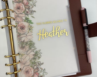 Puede incluir: Un planificador con un diseño floral y el texto dorado "THIS PLANNER BELONGS TO Heather". El planificador abierto revela un divisor transparente con un borde floral. Un bolígrafo con un barril de purpurina descansa sobre el planificador.