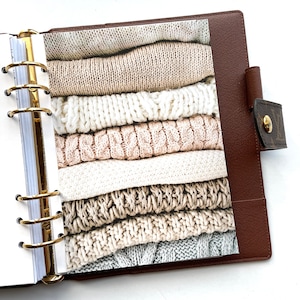 May include: A planner insert featuring a stack of folded sweaters in various neutral colors, including cream, beige, and gray. The sweaters have different knit patterns. The insert is inside a brown leather planner with gold rings.