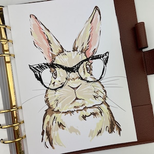 May include: A whimsical illustration of a rabbit wearing black-framed glasses. The rabbit is depicted in shades of brown, tan, and pink, with a sketched style. The image is on a white background, and is part of a planner.