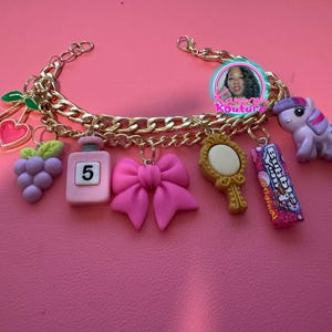 May include: A gold chain bracelet with multiple charms, including a pink bow, a purple bunch of grapes, a pink perfume bottle with the number "5" on it, a gold mirror, a purple unicorn, and a pink and blue bubble gum wrapper.