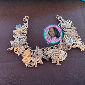 May include: Silver charm bracelet with various detailed charms, including a gingerbread house, ballet shoes, and a peace sign. The bracelet is displayed on a dark surface. A circular image with the text "By Dey" is in the background.