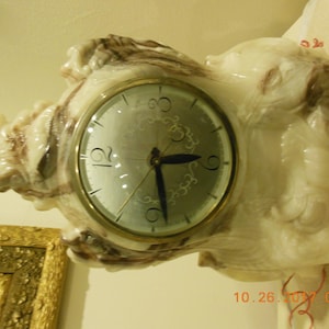 May include: A white and brown marble clock with a round face and black hands. The clock is in the shape of a stylized figure with a face and hands.