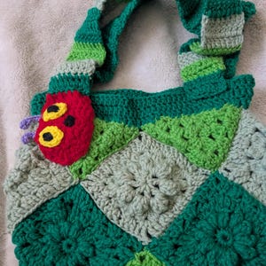 May include: A handmade crochet bag featuring a patchwork design in shades of green and gray. The bag has a green striped handle and a red caterpillar applique with yellow eyes. The bag is a unique, handcrafted accessory.