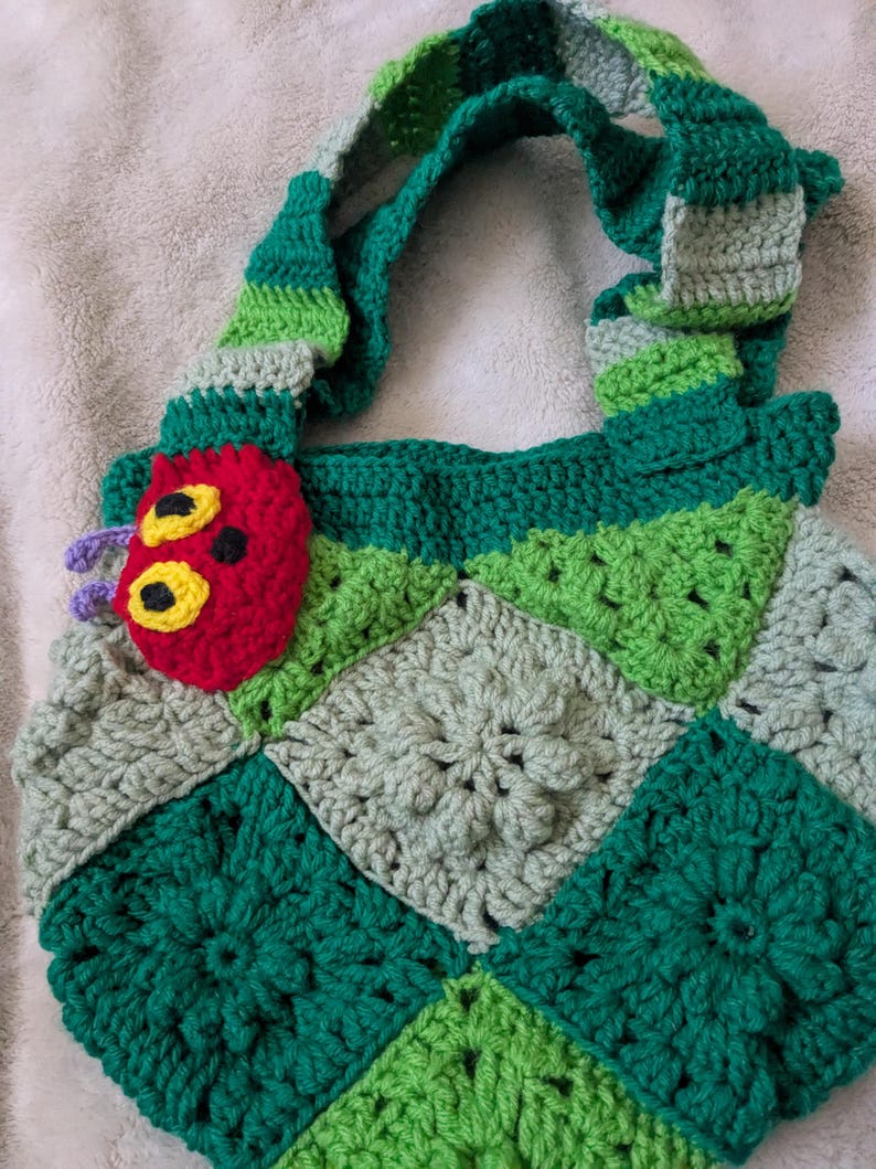 May include: A handmade crochet bag featuring a patchwork design in shades of green and gray. The bag has a green striped handle and a red caterpillar applique with yellow eyes. The bag is a unique, handcrafted accessory.