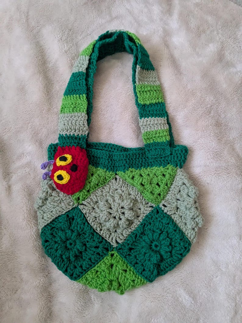 May include: A handmade crochet tote bag featuring a colorful design. The bag has a green, gray, and white striped handle and a patchwork body with green and gray squares. A red caterpillar applique with yellow eyes and purple antennae is attached to the side.