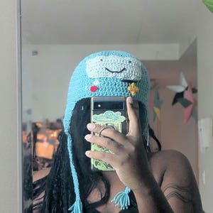May include: A light blue crocheted hat with ear flaps and braided tassels. The front features a white rectangular patch with a black embroidered smiling face, a small red circular accent, and a yellow four-pointed star shape stitched on.