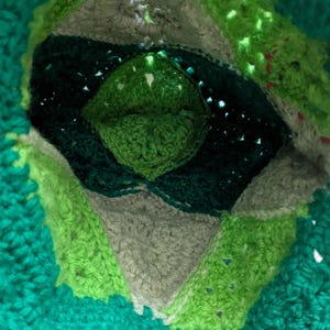 May include: Close-up of a handmade crochet item in shades of green, gray, and dark green. The design features a central diamond shape surrounded by concentric layers of varying textures and colors. The overall effect is a textured, layered design.