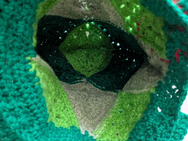 May include: Close-up of a handmade crochet item in shades of green, gray, and dark green. The design features a central diamond shape surrounded by concentric layers of varying textures and colors. The overall effect is a textured, layered design.