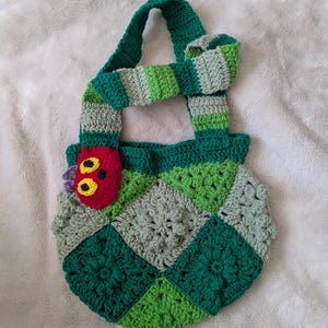 May include: A handmade crochet bag featuring a patchwork design in shades of green and gray. The bag has a green handle and a decorative caterpillar applique in red, yellow, and purple. The bag is approximately 10 inches wide.